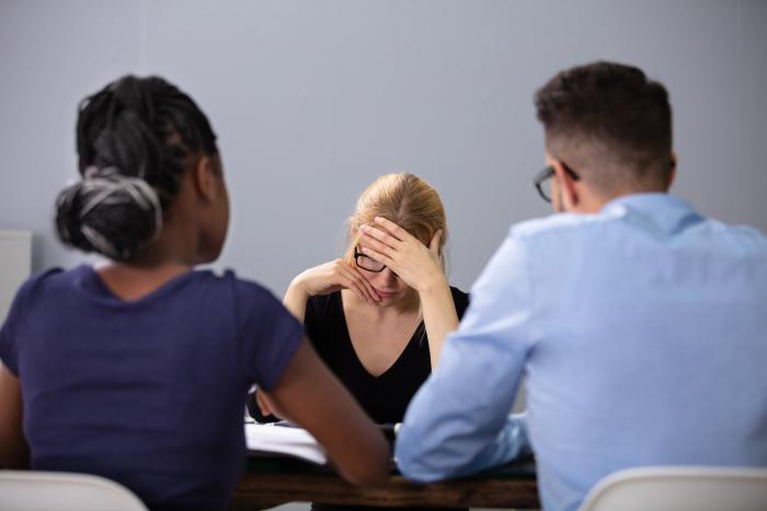 Three people sat at a desk, one looking despondent 