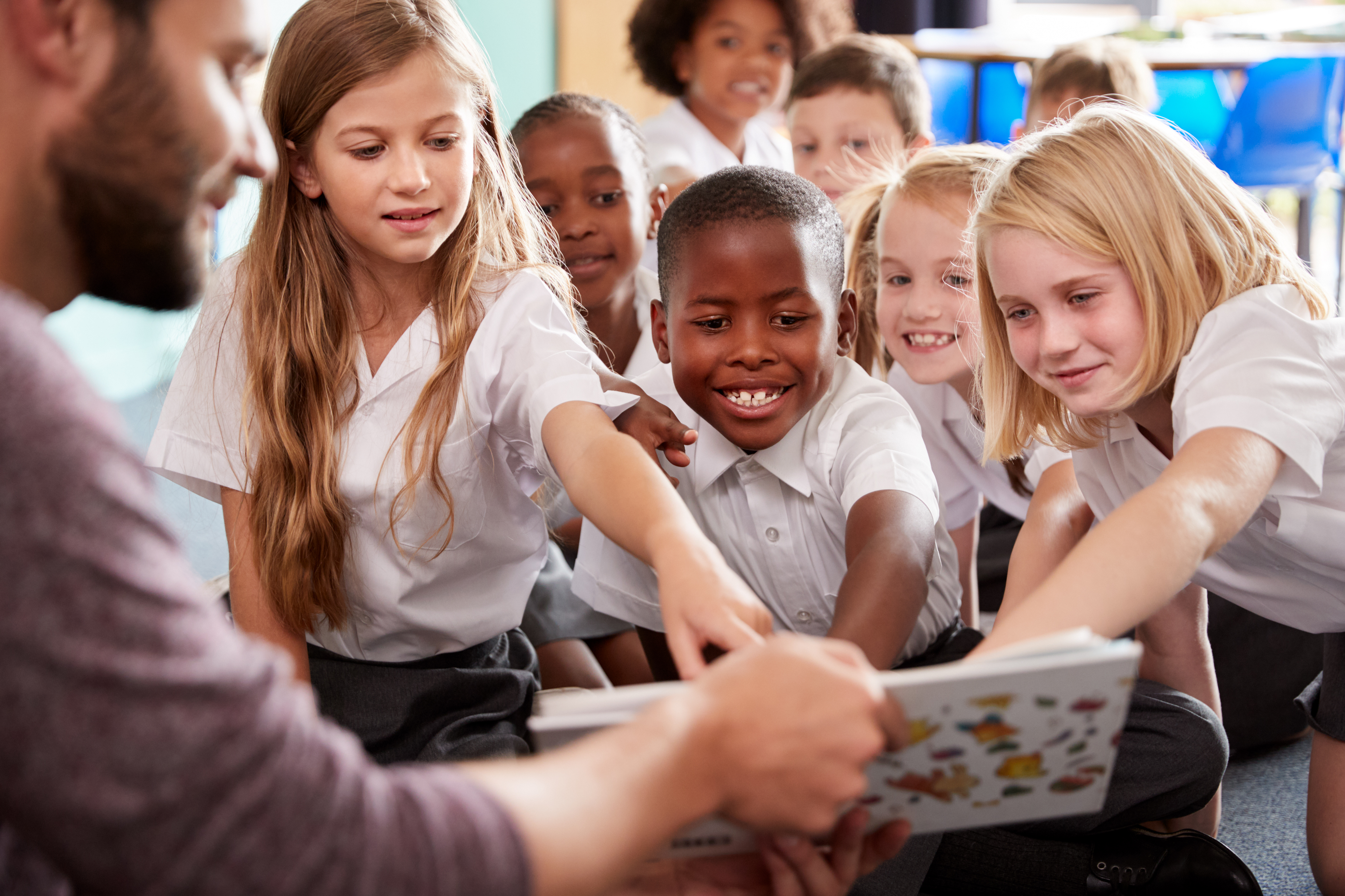 School children in white tops pointing into a book being held by an adult