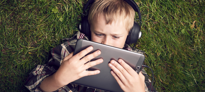 Child holding tablet close to face while laying on grass wearing headphones