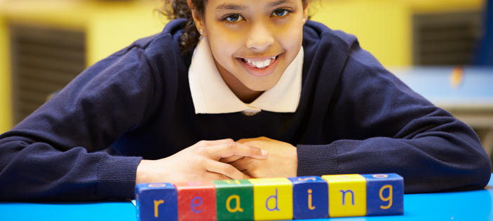 School girl in blue top smiling next to coloured word blocks which spell out "reading"