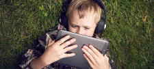 Child holding tablet close to face while laying on grass wearing headphones